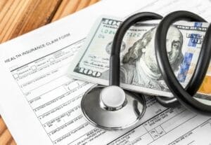 Stethoscope cord squeezing stack of $100 bills atop a health insurance claim for Wisconsin Medicaid