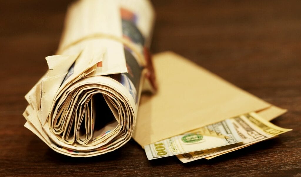 Rolled-up newspaper next to an envelope of U.S. currency bills, indicative of an alarming proposal for Wisconsin newspaper subsidies.