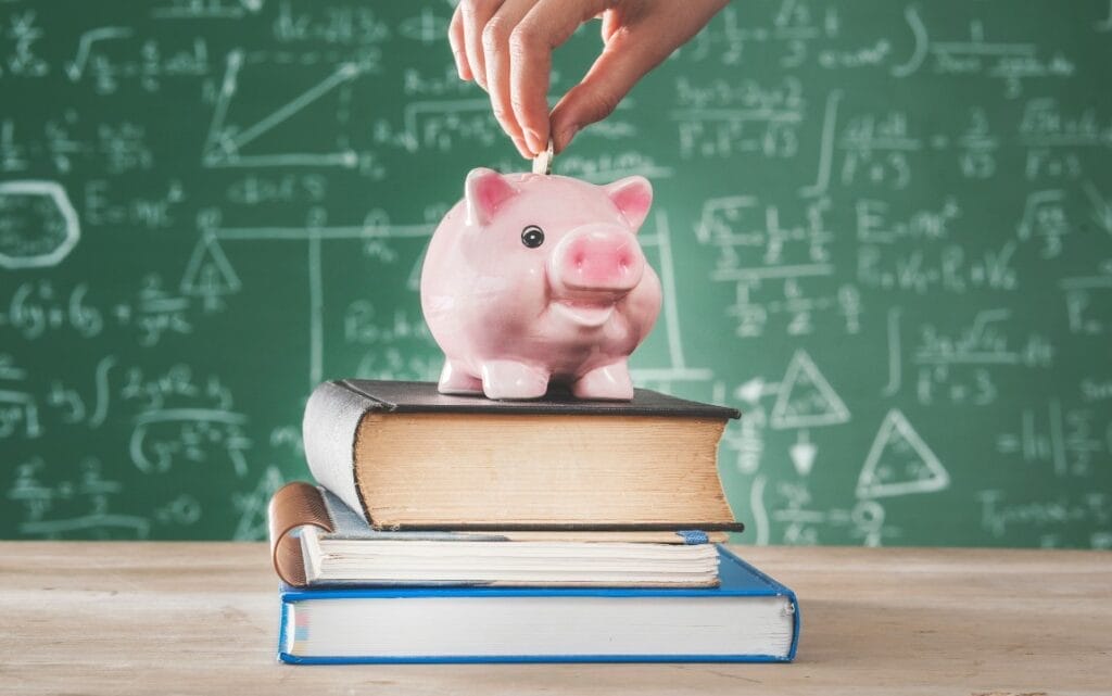 Person placing coin into piggybank set atop books and against a background of a school chalkboard.