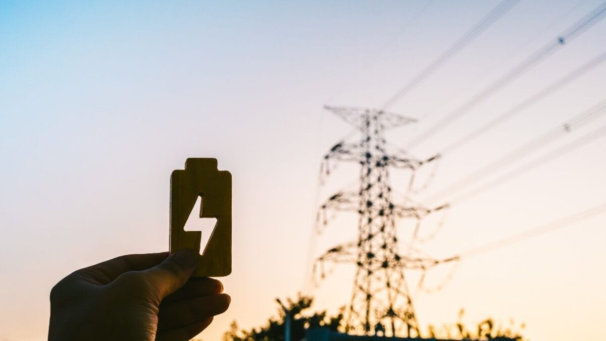 Hand holding a cutout of a battery icon against a backdrop of towering power lines