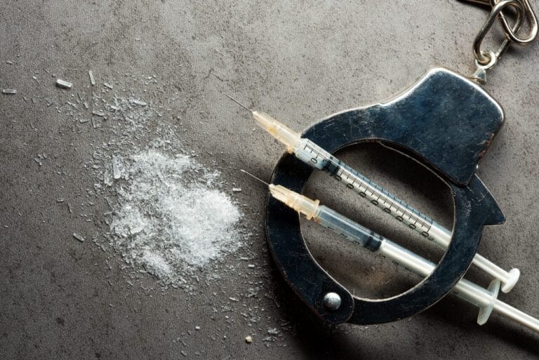 Image of handcuffs and drug-use syringes next to crystalized methamphetamine
